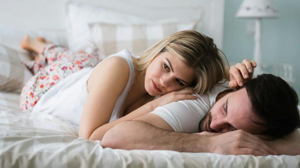 A woman lying on a bed with her husband, who is sleeping.
