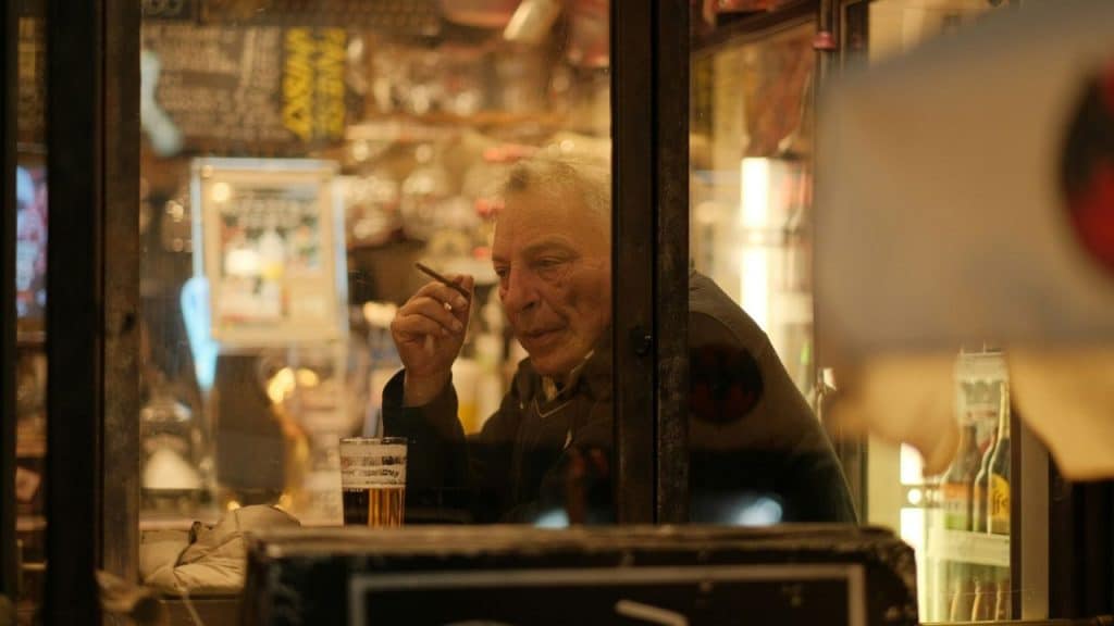 An older man with a cigarette sits behind a glass window with a glass of beer.