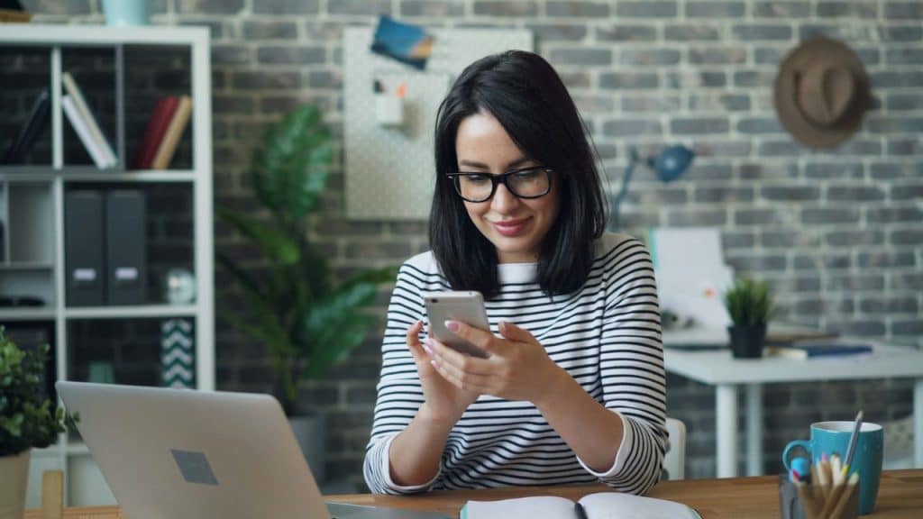 A woman wearing glasses and a striped shirt looks at a phone at a wooden desk.