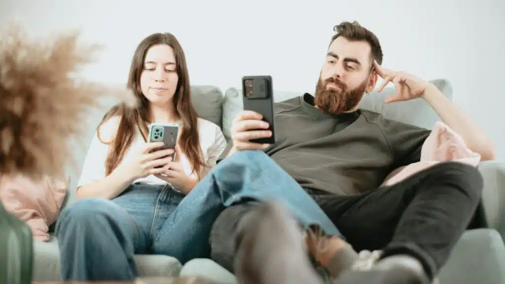 A man and woman sit on a couch, each looking at their own handheld smartphone.
