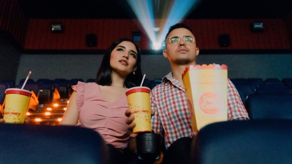 Two people in a theater look up at a light beam while holding large drinks.