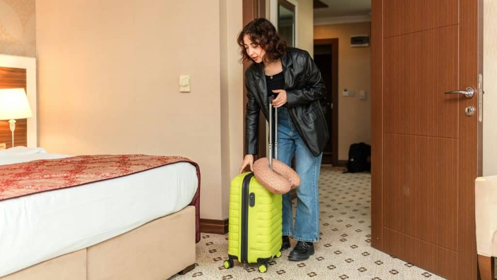 A woman in a leather jacket stands in a hotel room holding a bright green suitcase.
