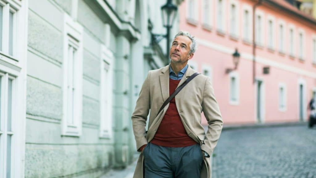 A man in a tan coat and red sweater walks down a European street.