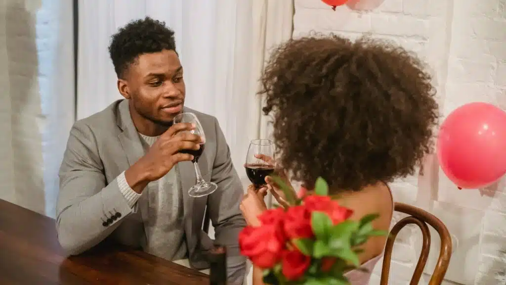 A man and woman sit at a table, holding wine glasses near red roses and balloons.
