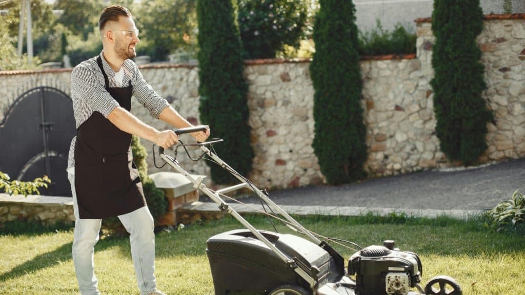 A man wearing a black apron and safety glasses pushes a lawnmower across green grass.
