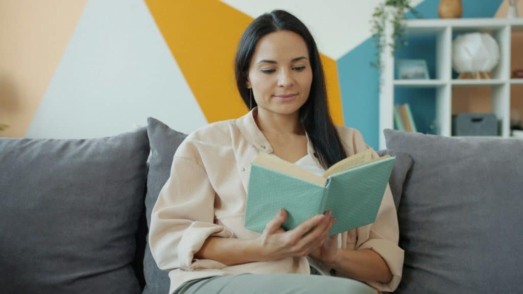 A woman with long dark hair sits on a gray sofa reading a teal book.