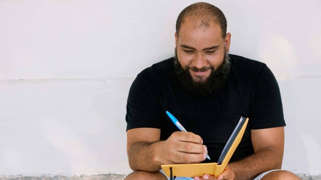 A man with a beard sits against a white wall and writes in a yellow notebook.