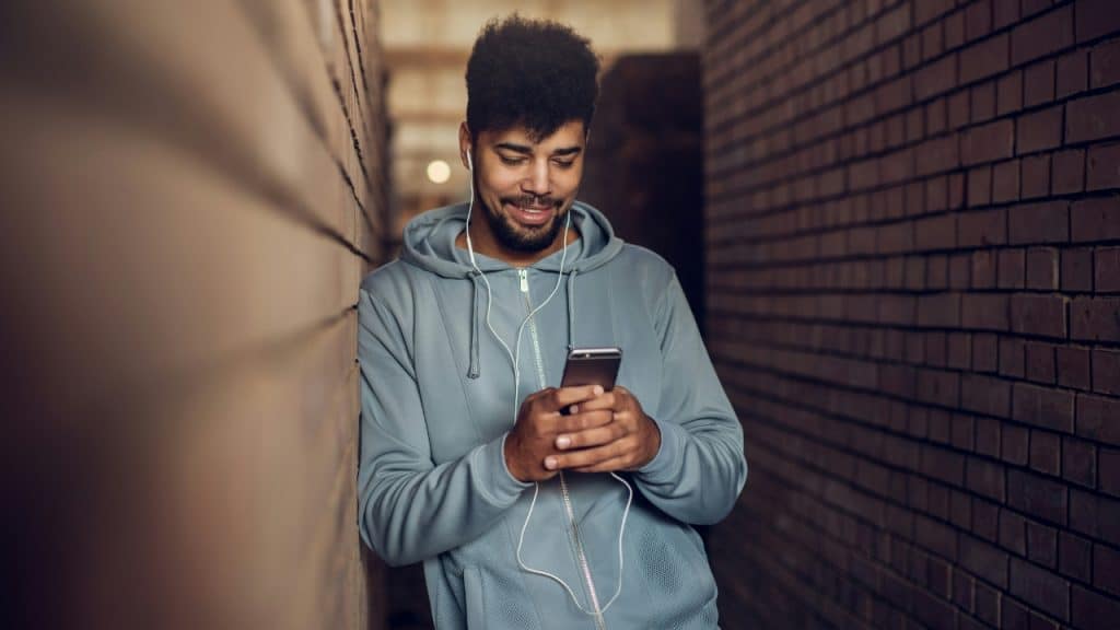 A man wearing a hoodie and earphones leans against a brick wall using a phone.