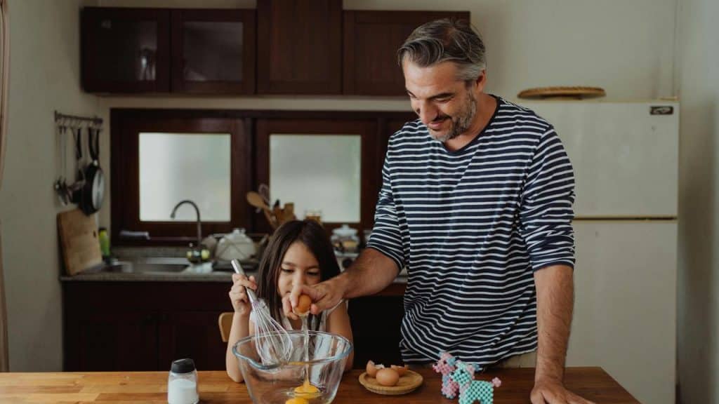 A man and a child cracking eggs together while preparing food in a kitchen.