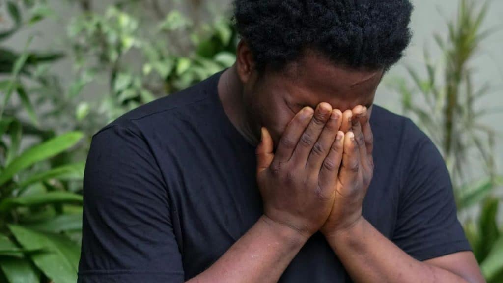 A person covering their face with both hands while standing outdoors.