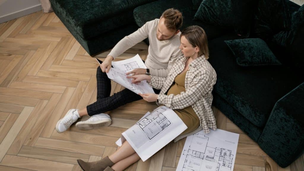 A man and a pregnant woman sit on a wooden floor reviewing architectural blueprints together indoors.