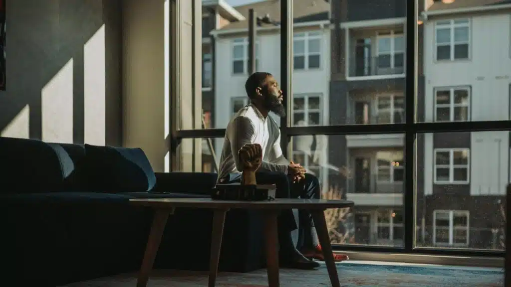 A bearded man sits on a dark sofa looking out a large window at apartment buildings.