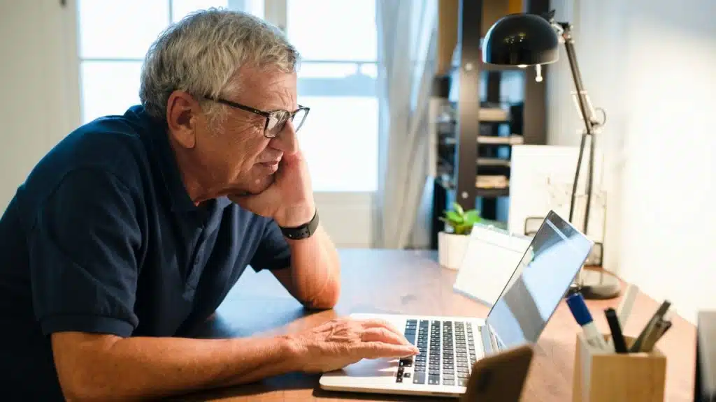An older man wearing glasses rests his chin on his hand while viewing a laptop.