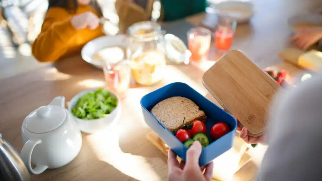Hands hold an open blue lunchbox containing a sandwich, cherry tomatoes, and cucumber slices.