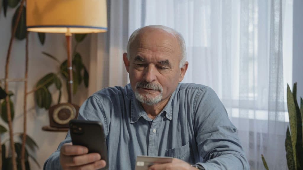 A man with a grey beard looks at his smartphone while holding a small plastic card.