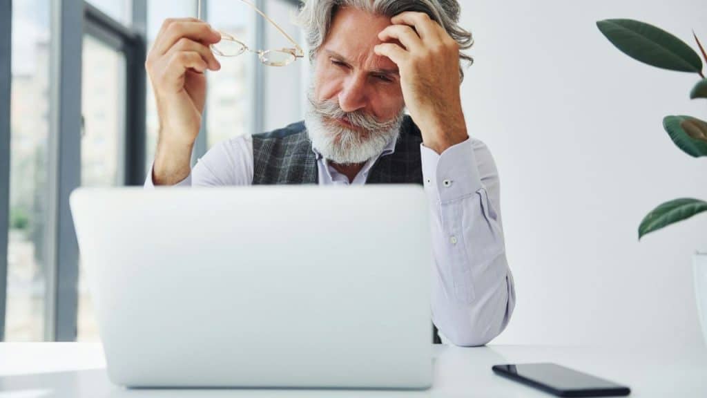 An older man with a gray beard sits at a laptop and holds his glasses tiredly.