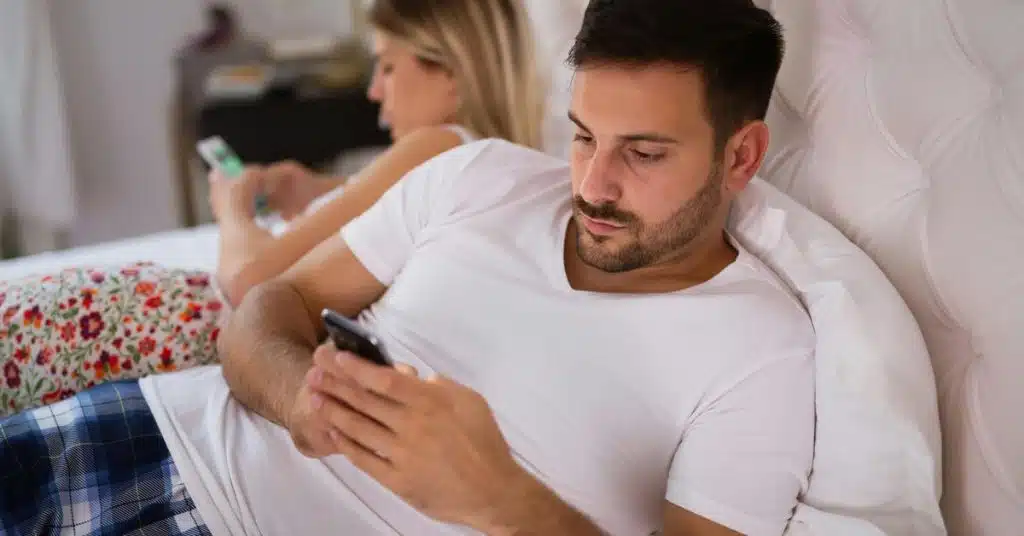 A couple lies on a bed while both look at their phones.