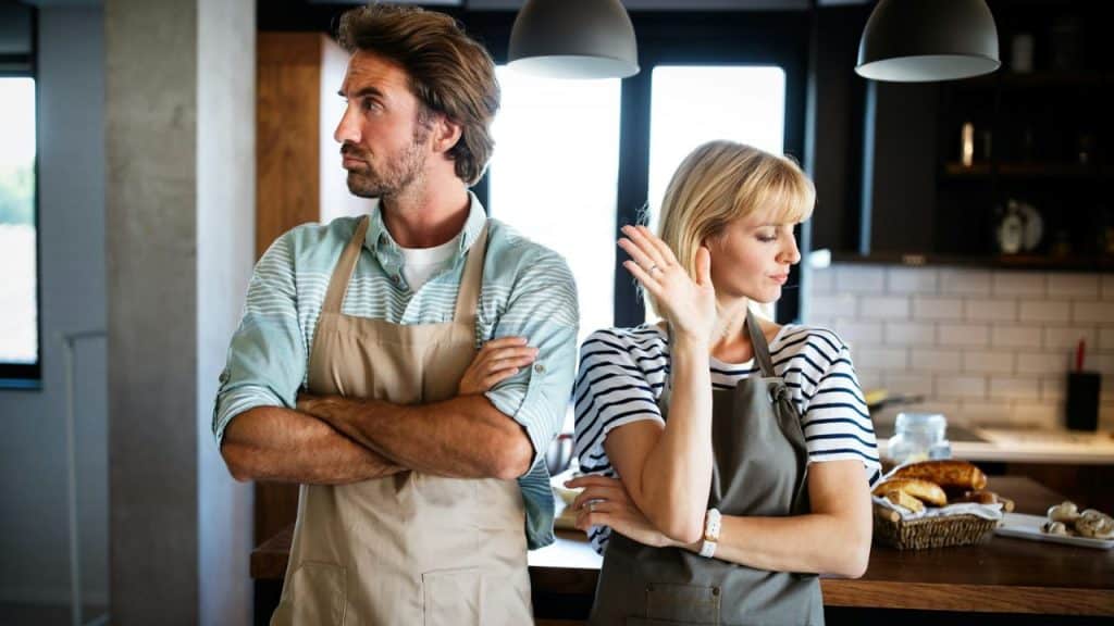 A man and a woman in aprons stand back-to-back in a kitchen with their arms crossed, looking annoyed.
