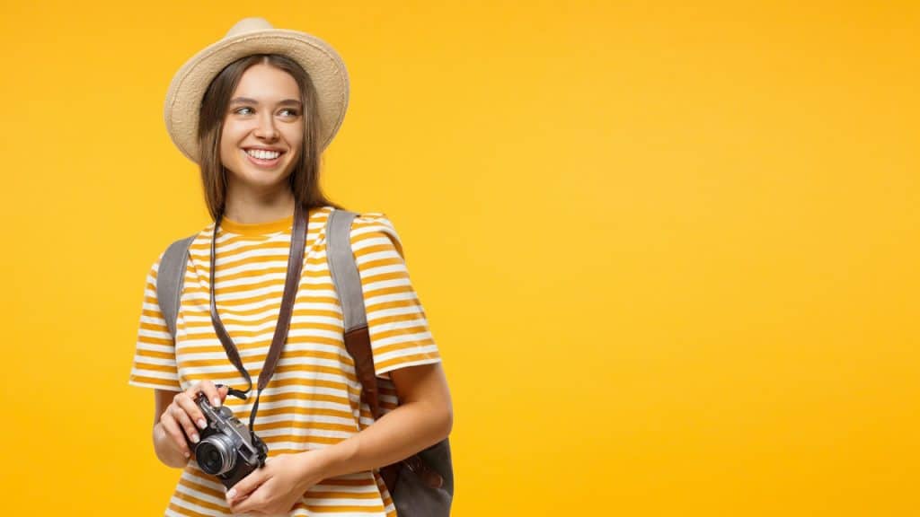 A man wearing a hat and holding a camera is smiling in front of a yellow background.