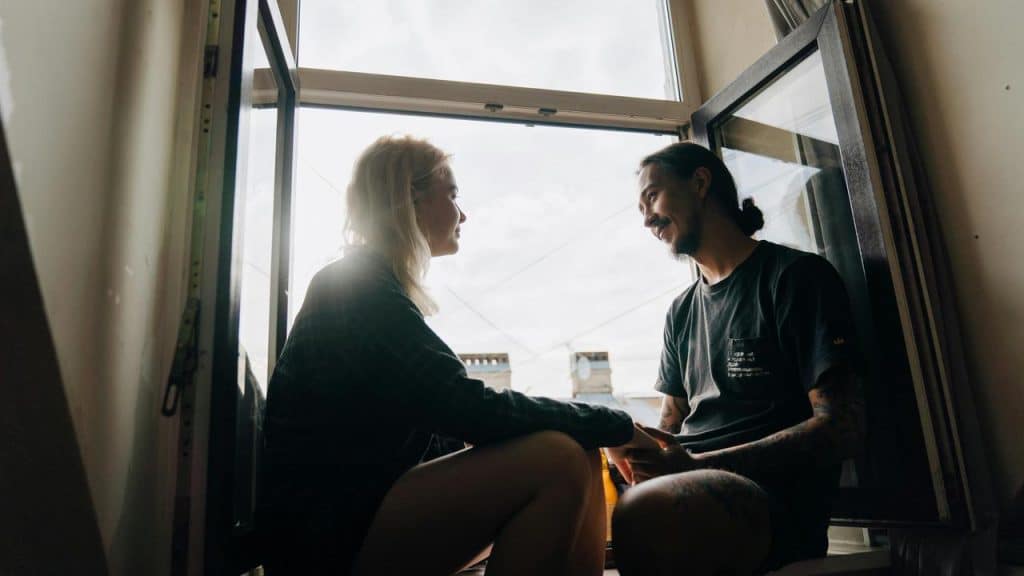 A couple sitting in an open window while smiling and talking.
