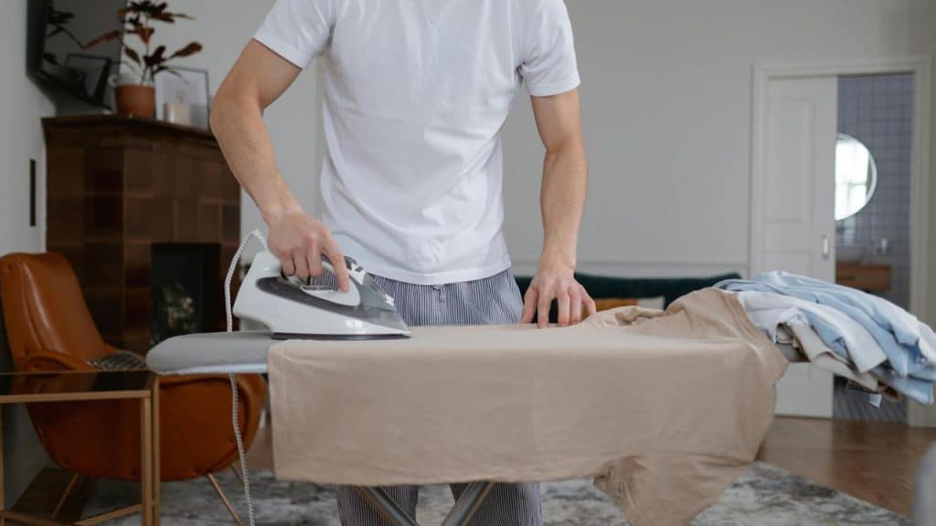 A person ironing a beige shirt on an ironing board in a tidy room.