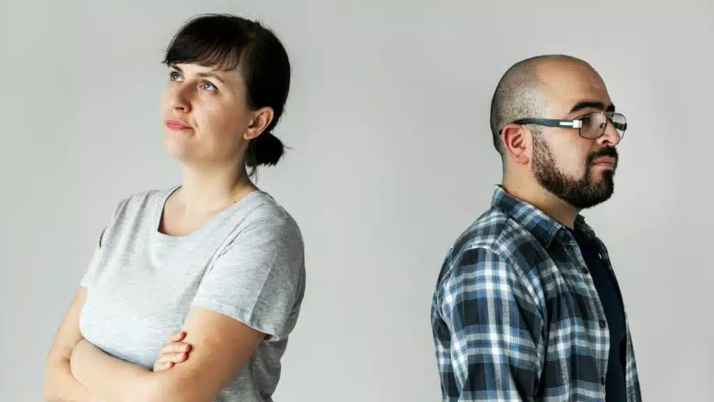 A woman and a man with glasses stand back-to-back against a plain gray wall.