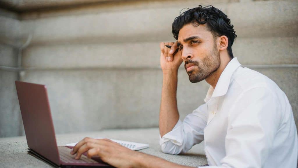 A man sitting outdoors with a laptop and looking thoughtful.