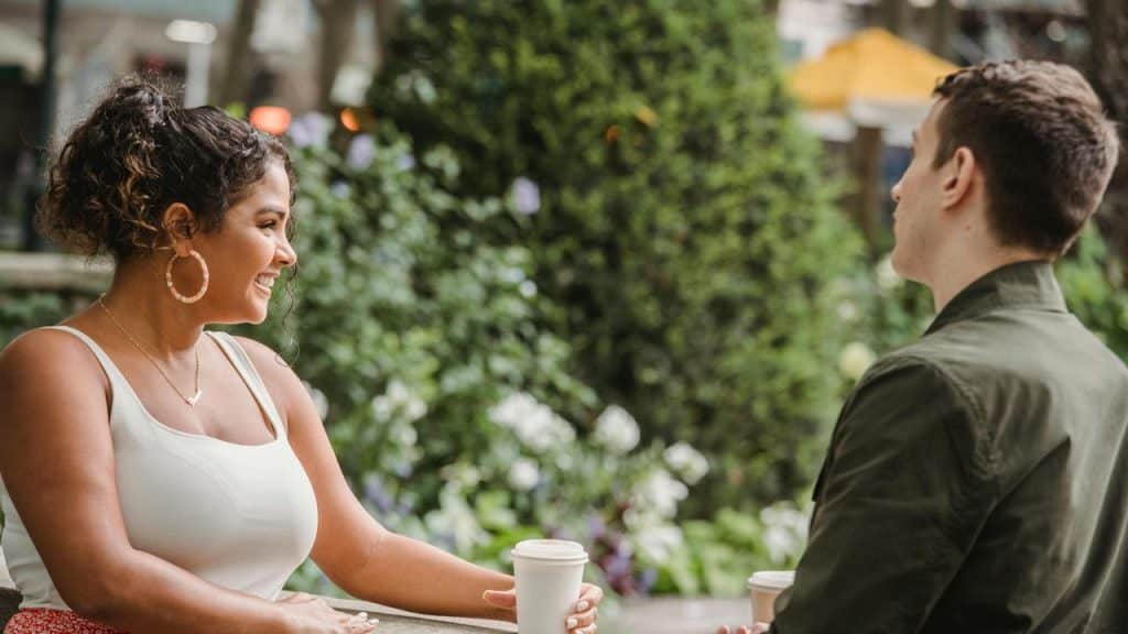 A woman smiling while talking to a man outdoors with coffee cups.