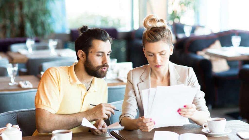 A man and woman sit at a table looking at papers together with serious facial expressions.