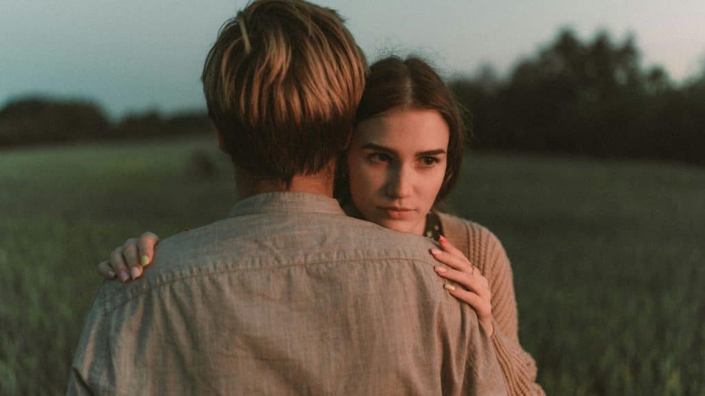 A woman rests her chin on a man's shoulder while looking away in a field.