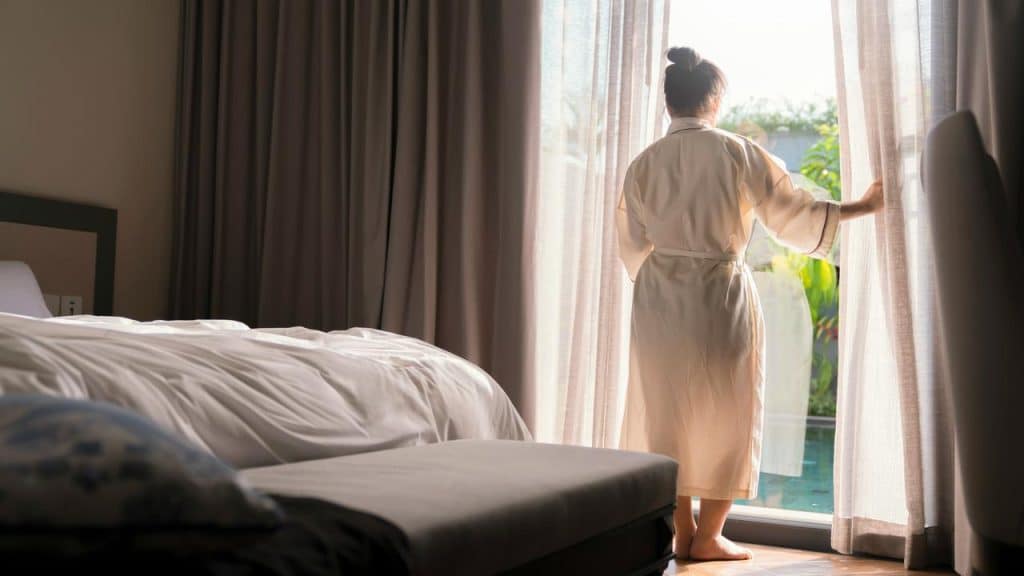 A woman in a robe standing by a window, looking outside.