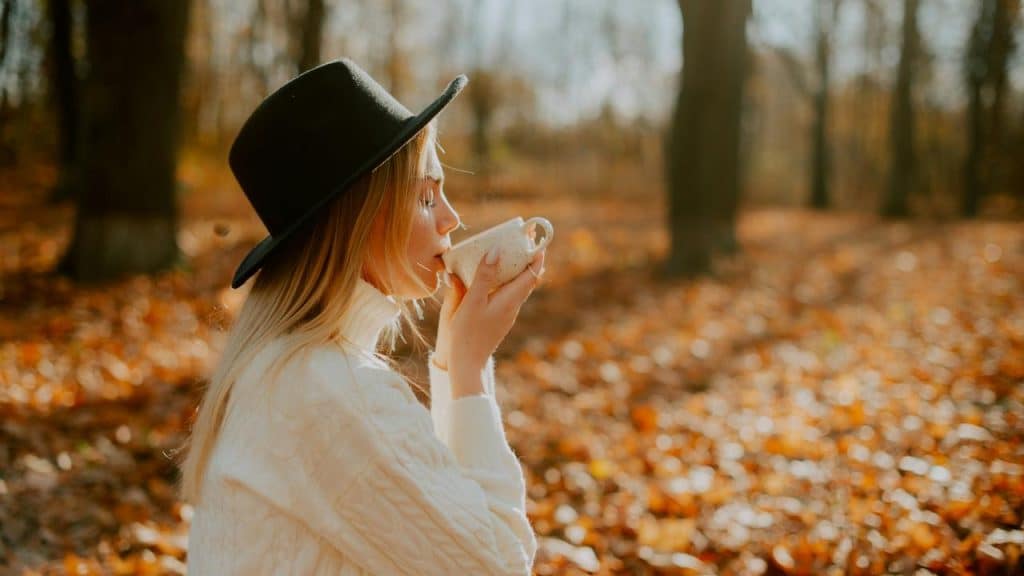 A person in a hat sips a warm drink in an autumn forest.
