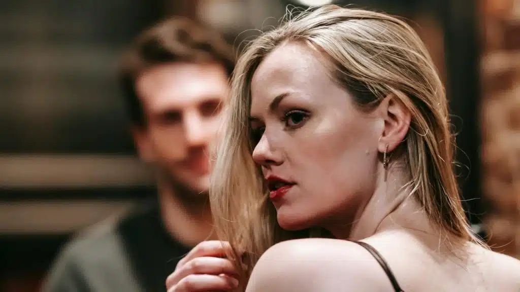 A blonde woman with red lipstick looks sideways with a blurred man in the background.