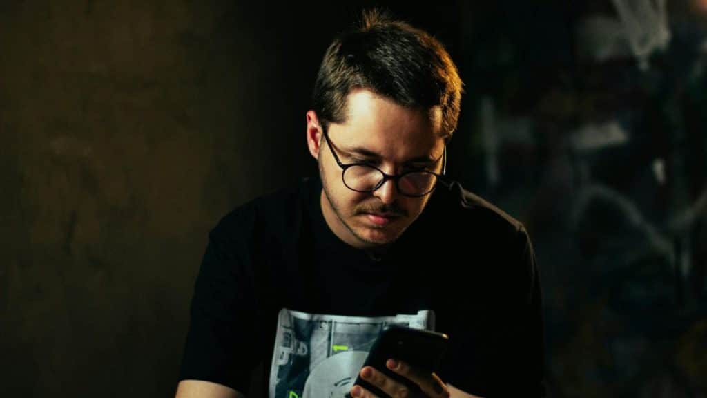 A man wearing glasses looks down at the screen of a smartphone in a dimly lit room.