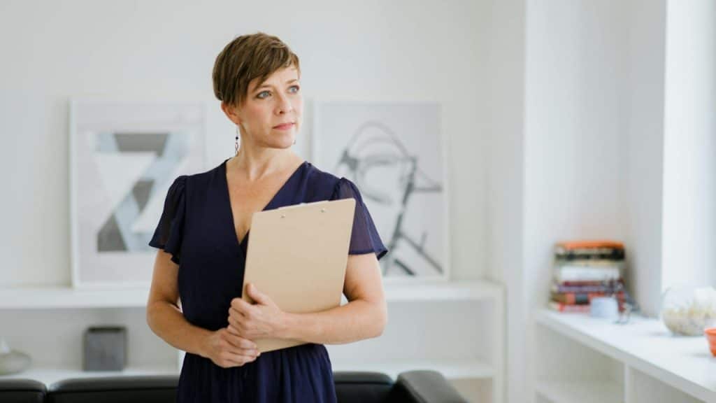 A woman with short hair and a blue dress holds a clipboard in an office.