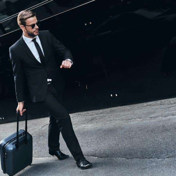 A young man in full suit pulling luggage while walking outdoors.