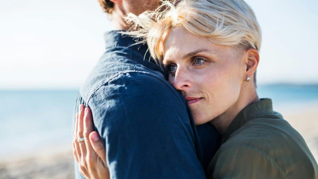 A woman hugging a partner while looking thoughtfully toward the sea.