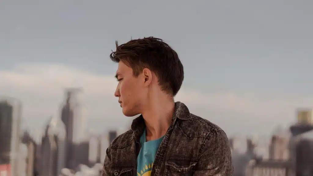 A short-haired person in a denim jacket looks sideways against a blurred city skyline.