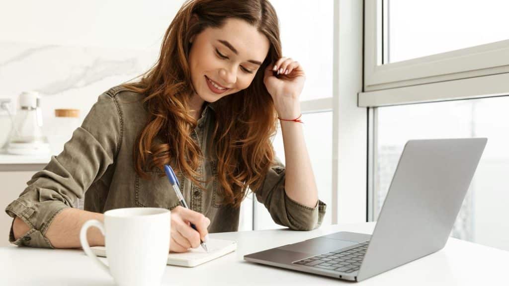 A woman smiling while writing in a notebook beside a laptop and coffee mug.