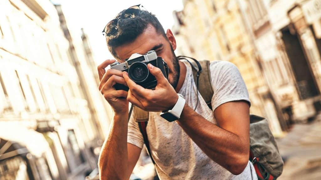 A man with a backpack holds a vintage camera to his eye on a city street.