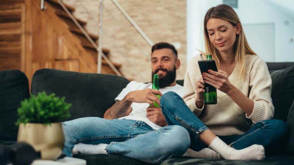 A man and woman sit on a sofa holding green bottles while looking at phones.