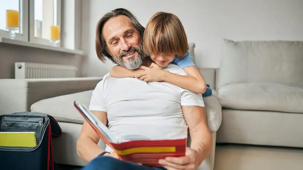 A child with his arms wrapped around his bearded, smiling father’s shoulders as he reads to him from a book while sitting on the floor and leaning against a couch.