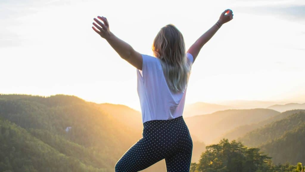 A woman is standing with outstretched arms on a cliff overlooking a picturesque view.