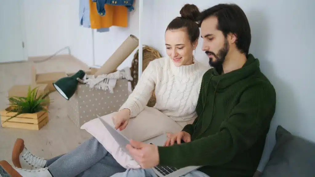 A couple sitting on the floor looking at a laptop near moving boxes.