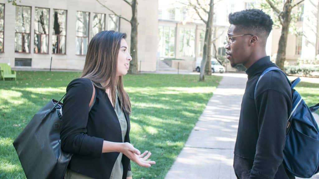A woman carrying a bag is talking to a bespectacled man who also carries a bag while they stand outdoors.