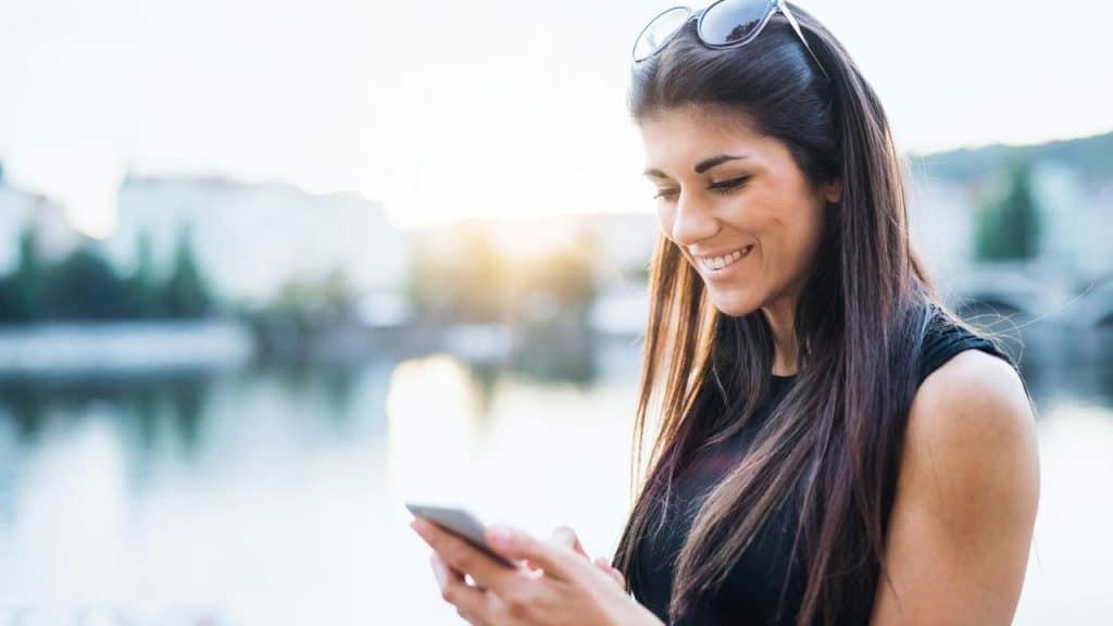 A woman smiling while looking at her phone outdoors near water.