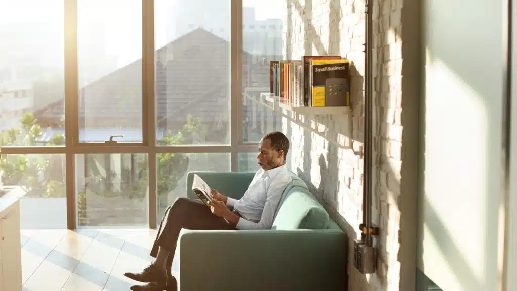 A man reads a book on a couch in a sunlit room.