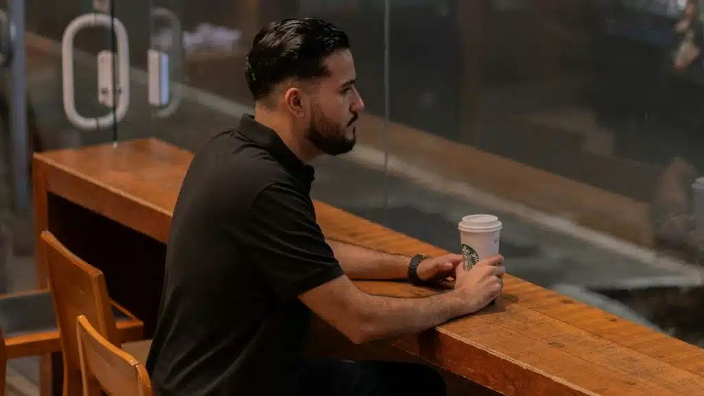 A man with a beard sits at a wooden counter in a cafe holding a white cup.