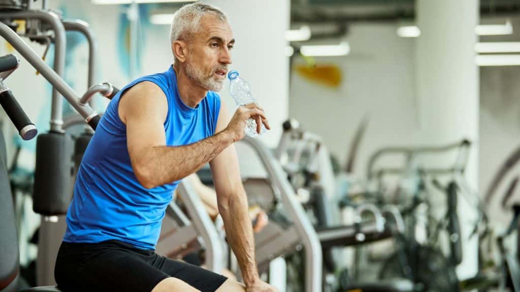 A man in a blue tank top sitting in a gym while drinking from a water bottle.