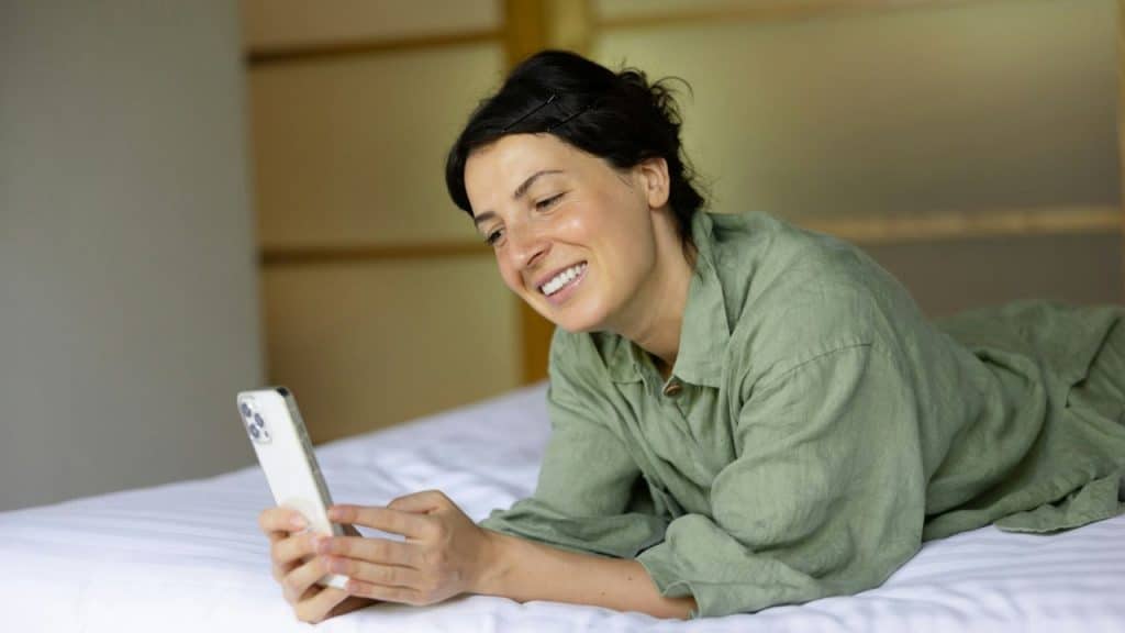 A smiling woman in a green shirt lies on a bed while holding a white smartphone.